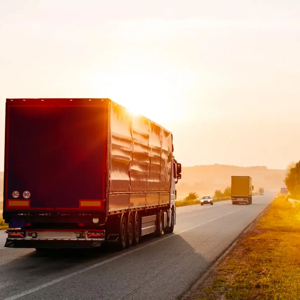 Camion che percorre un'autostrada al tramonto, con il sole che riflette sulla carrozzeria e altri veicoli in lontananza.